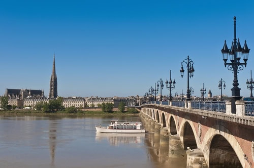 Blick auf die Saint Pierre Brücke in Bordeaux, im Hintergrund die Skyline der Stadt und im Vordergrund fährt ein Touristenschiff auf der Garonne